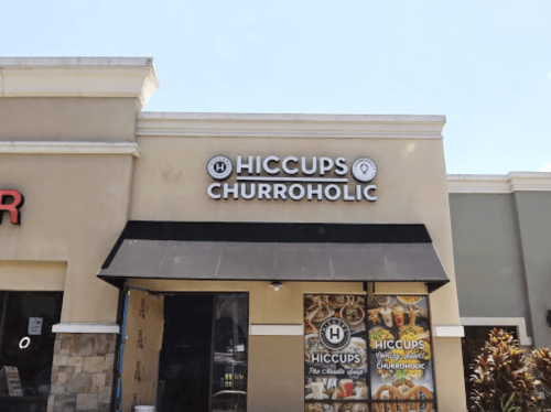 Sign for "Hiccups Churroholic" on a storefront, featuring a black awning and colorful window displays.