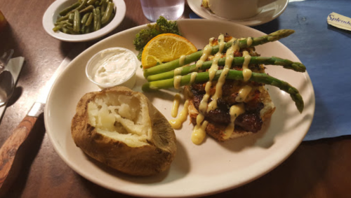 A plate with grilled chicken topped with asparagus, a baked potato, and a side of green beans and orange slices.