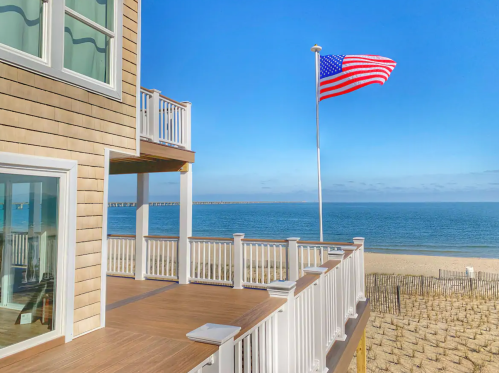 A beach house with a wooden deck and an American flag, overlooking a sandy beach and calm ocean under a clear blue sky.