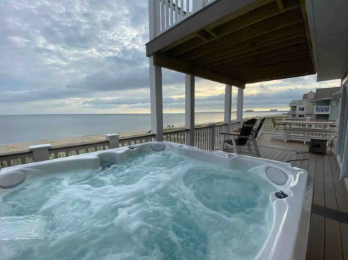 A hot tub on a deck overlooking a beach at sunset, with calm waves and a cloudy sky in the background.