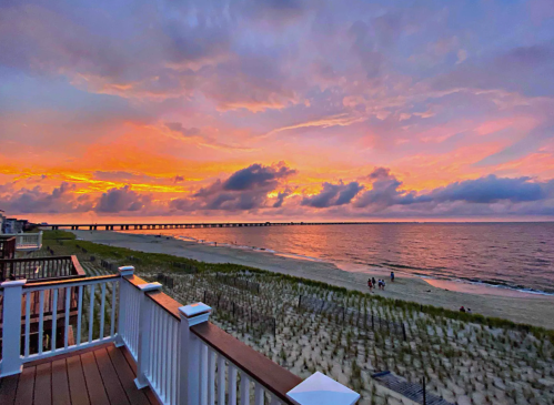 A vibrant sunset over the ocean, with clouds reflecting orange and purple hues, viewed from a beachside deck.