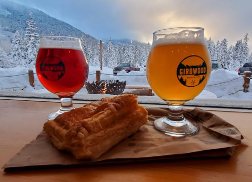 Two glasses of beer, one amber and one golden, sit on a table with a pastry, against a snowy mountain backdrop.