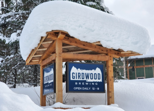 Sign for Girdwood Brewing, covered in snow, with hours displayed: "Open Daily 12-8." Surrounded by winter scenery.