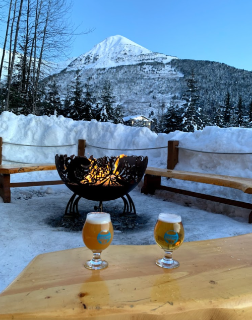Two glasses of beer sit on a wooden table near a fire pit, with a snowy mountain backdrop under a clear blue sky.