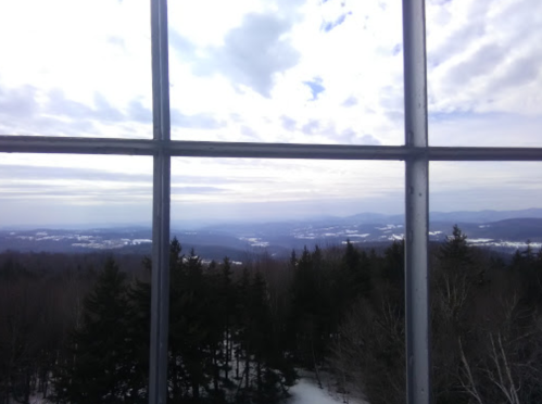 View from a tower window overlooking a snowy landscape with mountains and a cloudy sky in the distance.