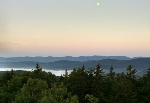 A serene mountain landscape at dusk, with a pale moon and mist rising over the distant hills.