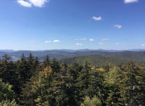 A panoramic view of rolling mountains and lush green trees under a clear blue sky with scattered clouds.