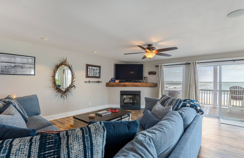 Cozy living room with a blue sofa, fireplace, TV, and large windows overlooking the beach. Bright and inviting atmosphere.