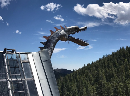 Metal dragon sculpture atop a roof, overlooking a forested landscape under a blue sky with clouds.