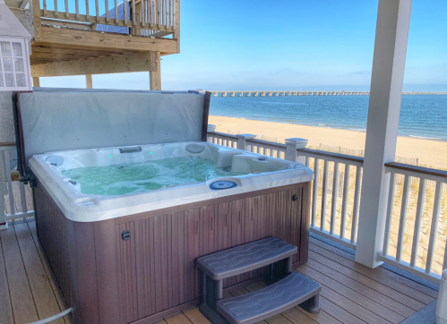 A hot tub on a deck overlooking a beach and pier, with clear blue skies in the background.