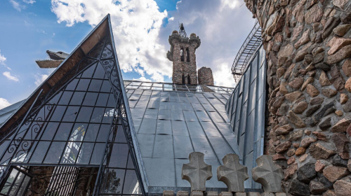 A unique architectural structure featuring a stone tower, glass elements, and a sloped metal roof against a cloudy sky.