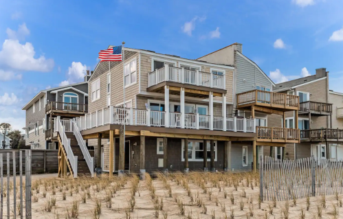 A beach house with multiple decks, an American flag, and sandy dunes in the foreground under a blue sky.