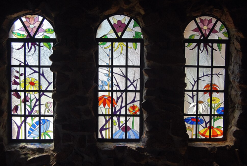 Three arched stained glass windows featuring colorful floral designs against a stone wall backdrop.