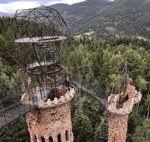 A stone tower with a metal dome and spiral staircase, surrounded by lush green trees and mountains in the background.