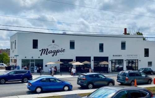A white building with "Magpie" signage, featuring outdoor seating and cars parked in front on a sunny day.