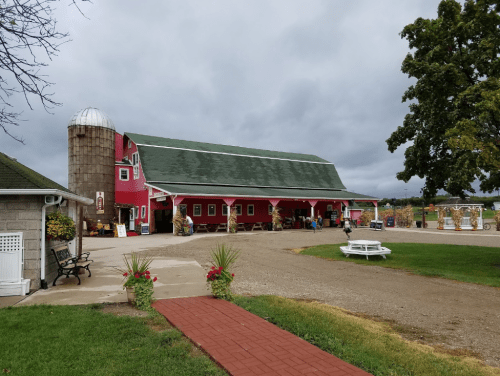 A red barn with a silo, surrounded by a gravel path, benches, and greenery under a cloudy sky.