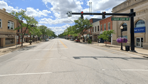 A quiet street view with shops and trees under a partly cloudy sky, featuring a traffic signal and street sign.