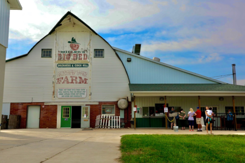 A farm building with a large sign, featuring visitors lined up outside on a sunny day.