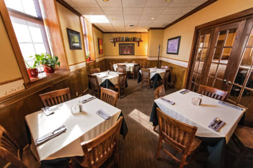 Cozy restaurant interior with wooden tables, chairs, and natural light, featuring neatly set tables and warm decor.
