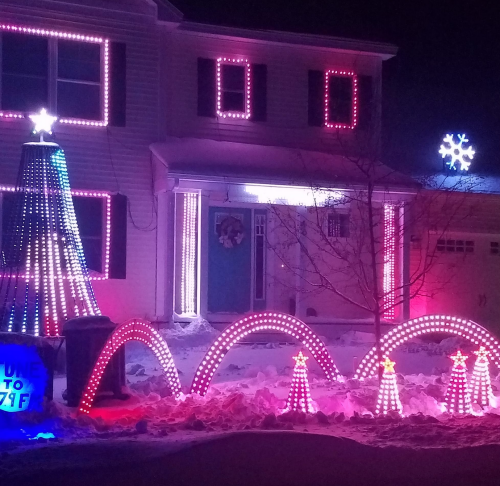 A house decorated with colorful holiday lights, featuring a lit tree, stars, and a snowflake, surrounded by snow.