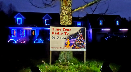 A sign on a tree reads "Tune Your Radio To 91.7 FM" in front of a house decorated with colorful lights.