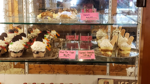 Display case filled with various desserts, including cupcakes, pie cups, and caramel cheesecake, with price tags.