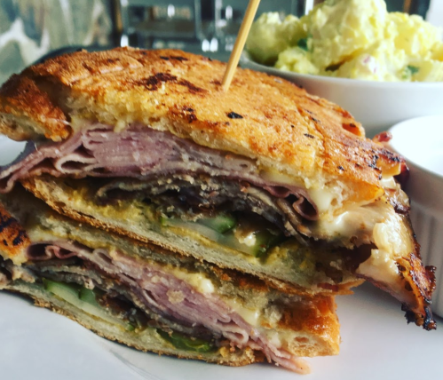 A close-up of a toasted sandwich filled with layers of meat and cheese, served with a side of potato salad.
