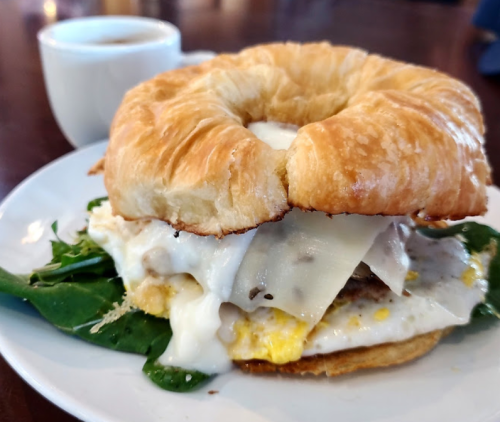A croissant sandwich filled with spinach, egg, cheese, and a side of coffee on a wooden table.