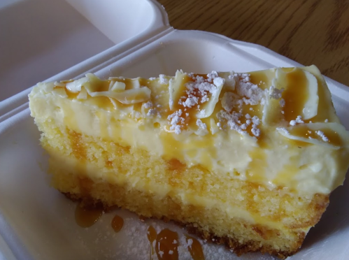 A slice of creamy cake with a yellow sponge base, topped with white frosting and drizzled with caramel sauce.