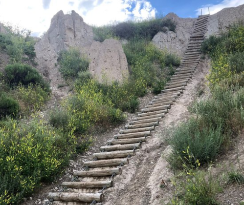 A wooden staircase leads up a hillside covered in green vegetation and rocky terrain under a cloudy sky.