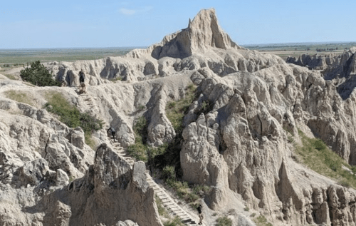 A rugged landscape with steep rock formations and a winding staircase leading through the Badlands under a clear blue sky.