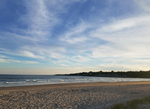 A serene beach scene at sunset, with gentle waves and a clear blue sky dotted with clouds.