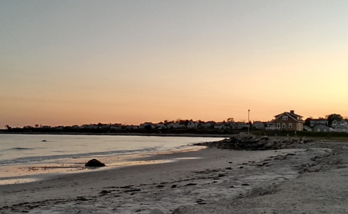 A serene beach at sunset, with gentle waves and a distant shoreline lined with houses under a pastel sky.