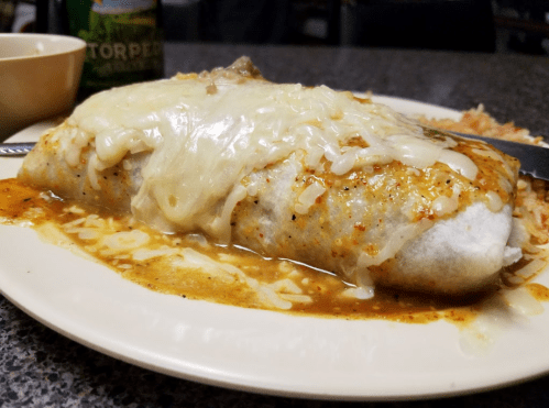 A large burrito topped with melted cheese and sauce, served on a plate with rice and a drink in the background.