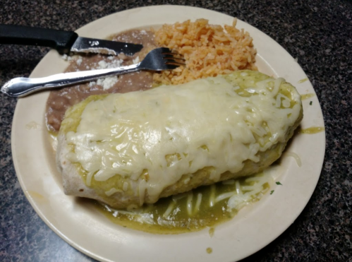 A plate with a large burrito covered in cheese and green sauce, served with rice and refried beans.