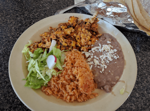 A plate of Mexican food featuring scrambled eggs, rice, refried beans, lettuce, and a radish garnish.