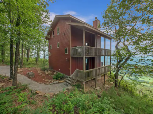 A red wooden cabin surrounded by trees, featuring a balcony and stone pathway leading to the entrance.