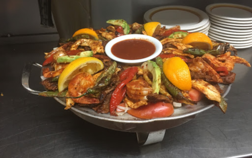 A large platter of grilled chicken, shrimp, and vegetables, garnished with lemon and orange slices, served with dipping sauce.