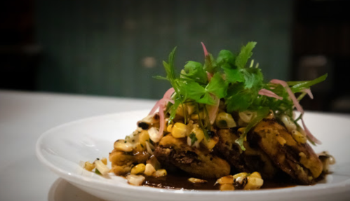 A plate of grilled plantains topped with corn, herbs, and pickled onions, served on a white dish.