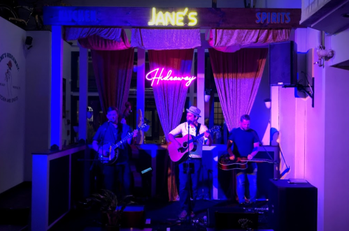 A band performs on stage at Jane's, with neon lights and curtains in a cozy venue setting.