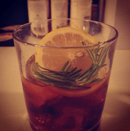 A glass of cocktail with a lemon slice and rosemary garnish, set against a blurred background of bottles.