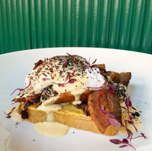 A plate of toast topped with poached egg, creamy sauce, crispy toppings, and microgreens, set against a green background.