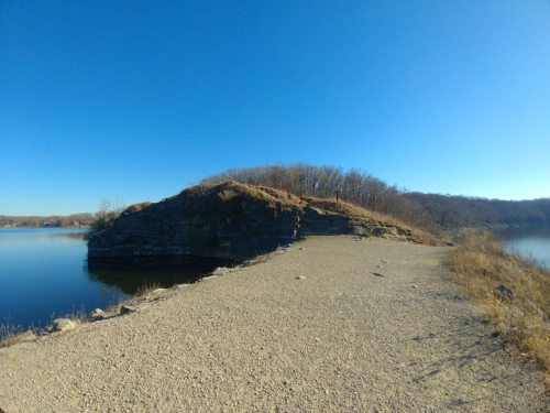 A rocky shoreline with a grassy hill, surrounded by calm water under a clear blue sky.