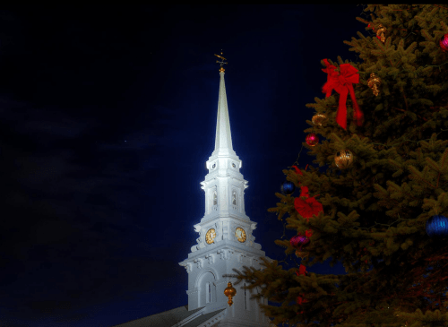 A church steeple illuminated at night, with a decorated Christmas tree in the foreground.