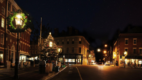 A charming street at night, adorned with festive lights and decorations, including a Christmas tree and wreaths.