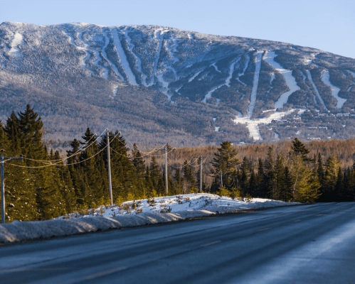 A clear road leads towards a snow-covered mountain with ski trails, surrounded by trees in a winter landscape.
