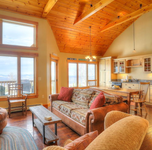 Cozy living room with wooden beams, large windows, and a view, featuring a sofa, chairs, and a dining area.