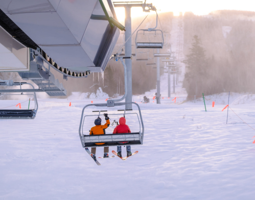 Two skiers on a chairlift, surrounded by snowy slopes and trees, with a soft sunset glow in the background.
