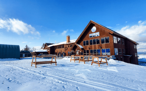 A wooden lodge with large windows sits on snowy terrain under a blue sky, with people gathered outside.