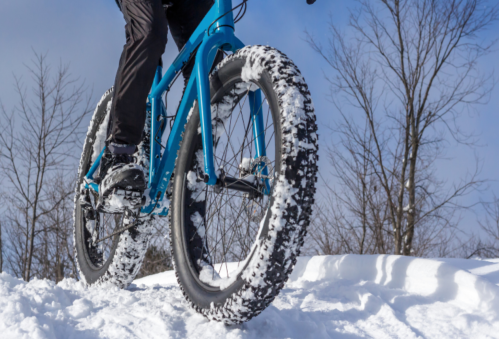 A cyclist rides a blue fat bike through snowy terrain, with large, snow-covered tires and bare trees in the background.